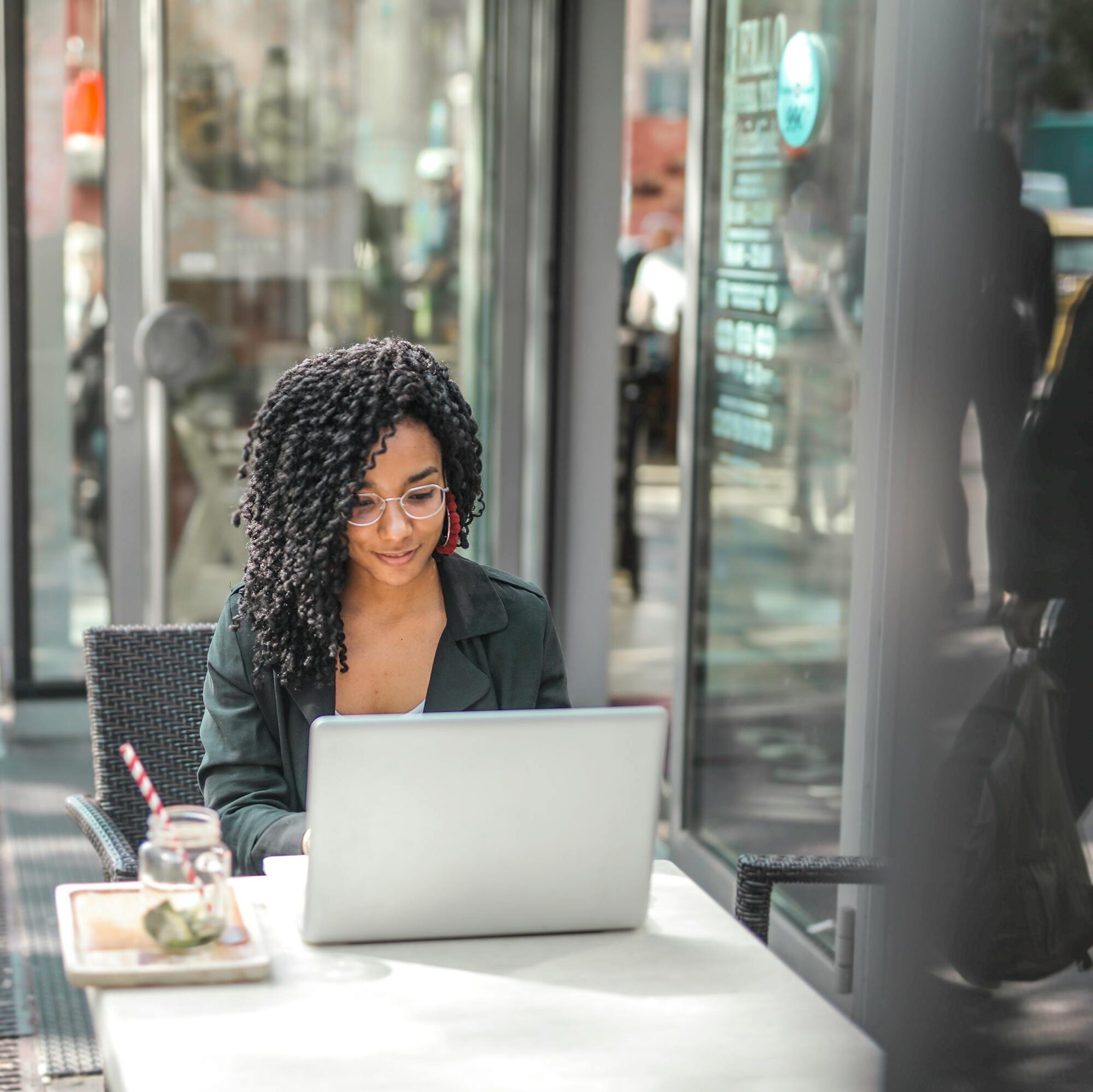 High angle of pensive African American female freelancer in glasses and casual clothes focusing on screen and interacting with netbook while sitting at table with glass of yummy drink on cafe terrace in sunny day